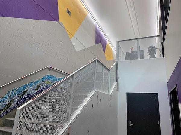 stairway leading up with metal mesh railings. the top of the wall is painted in swaths of purple, gold, and white, a mural runs along the wall side going up the stairs