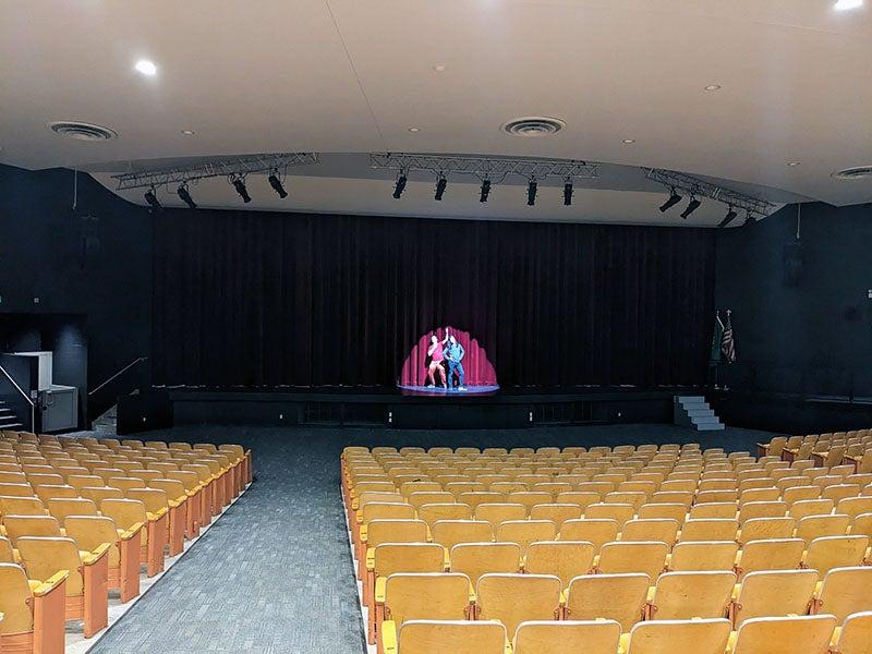 一个有浅棕色座位的礼堂面向舞台，舞台上有两个人在聚光灯下