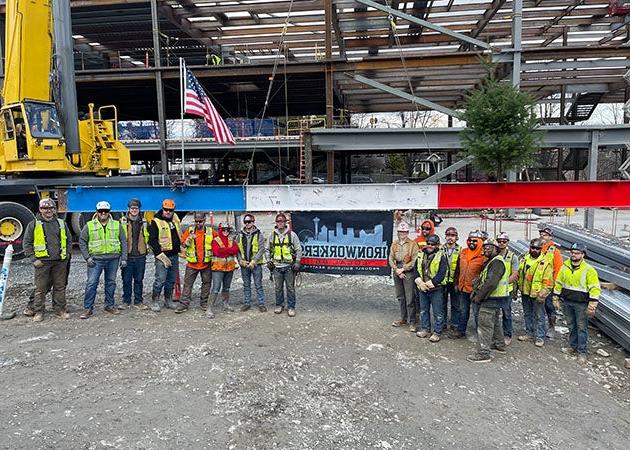 a group of people in construction safety vests and hats stand in front of a steel beam with a steel framed structure behind